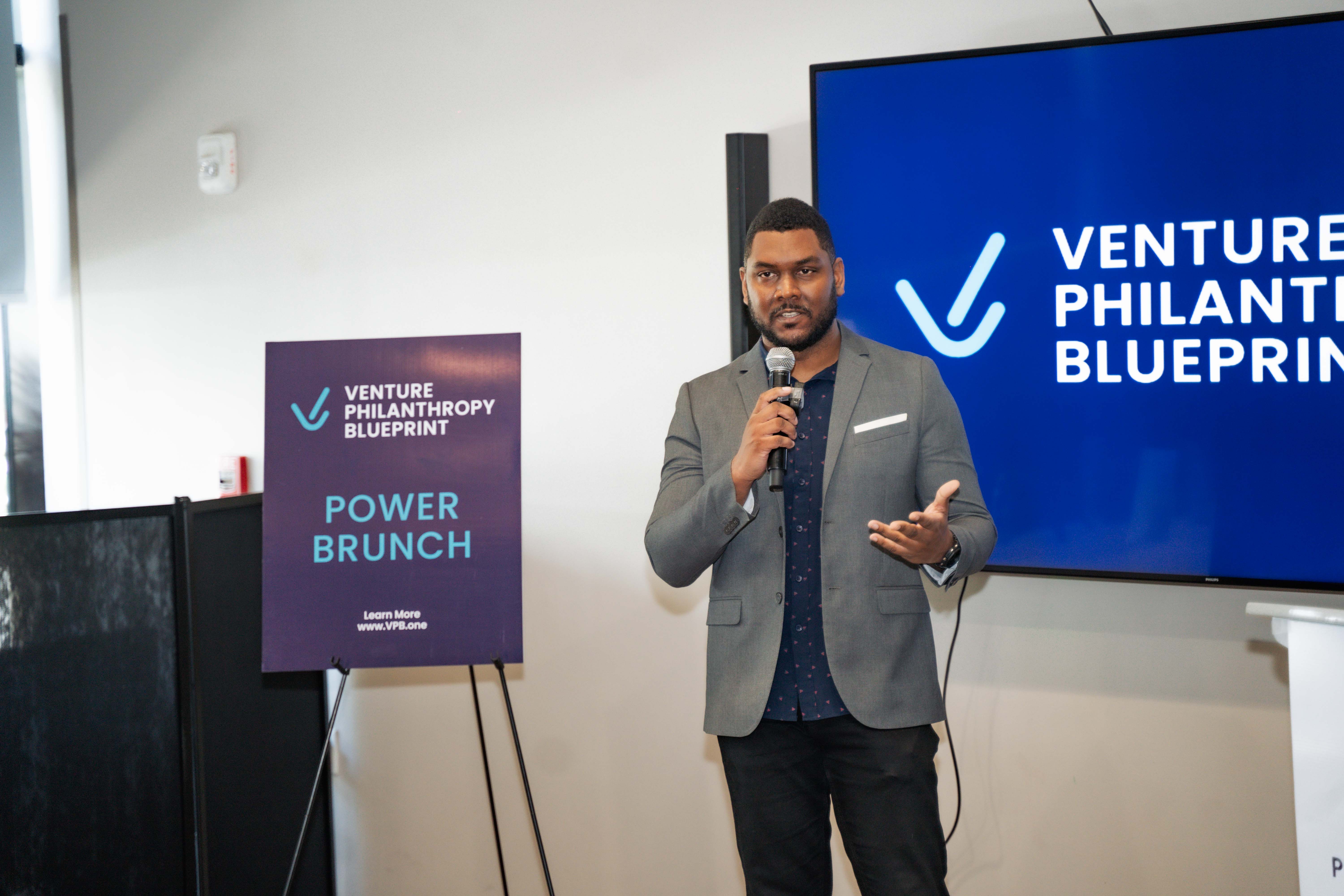 Paul Stennett speaks into a microphone at the Venture Philanthropy Blueprint Power Brunch event in Kingston, Jamaica. He stands in front of a screen displaying the event’s branding and beside a sign that reads “Power Brunch,” engaging the audience in a professional and inspiring setting focused on social impact and innovation.
