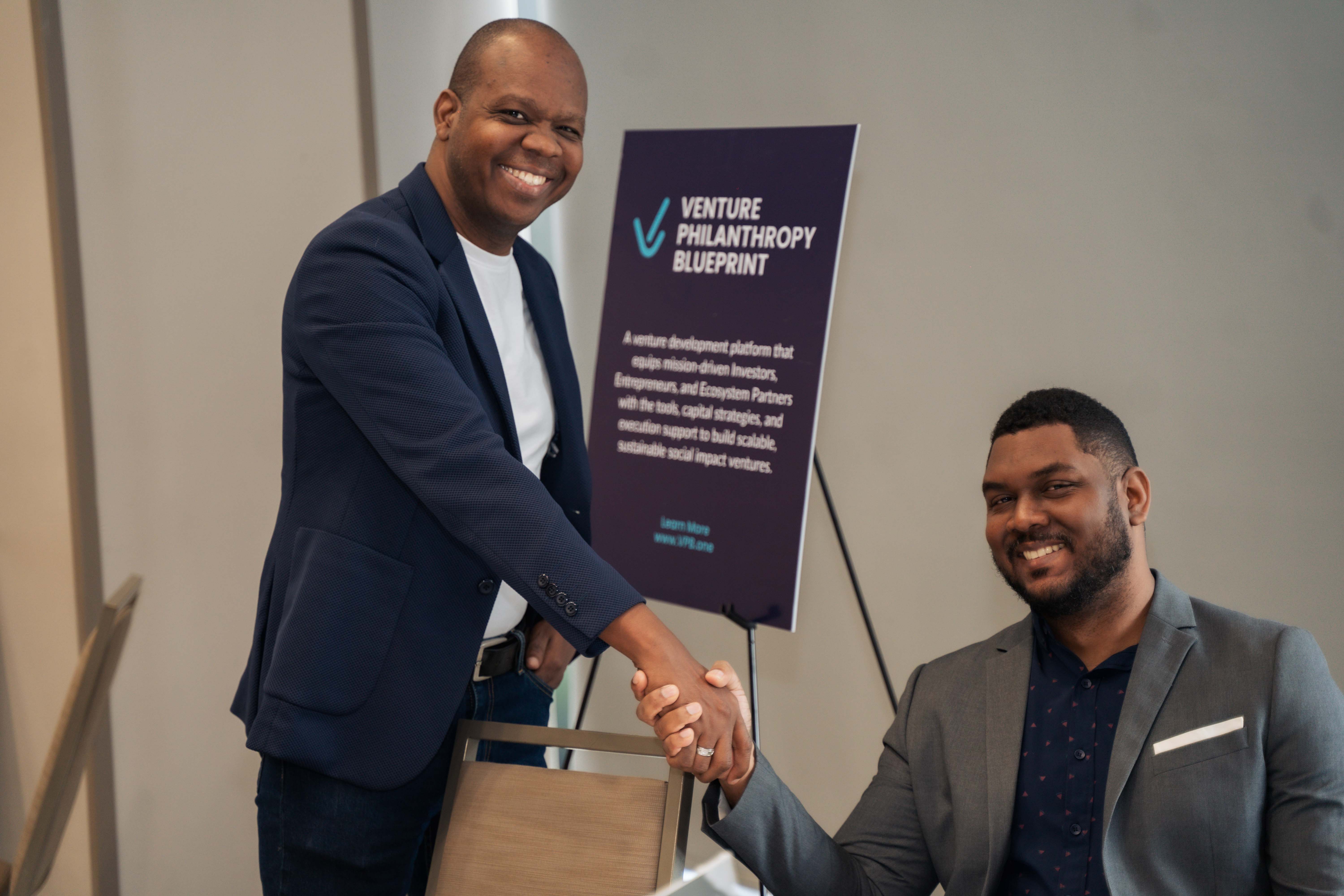 Almando Cox (left) shakes hands and smiles with Paul Stennett (right) at the Venture Philanthropy Blueprint Power Brunch event in Kingston, Jamaica. They are standing beside a branded sign highlighting the mission of the VPB platform, symbolizing collaboration and shared commitment to social impact and venture development.