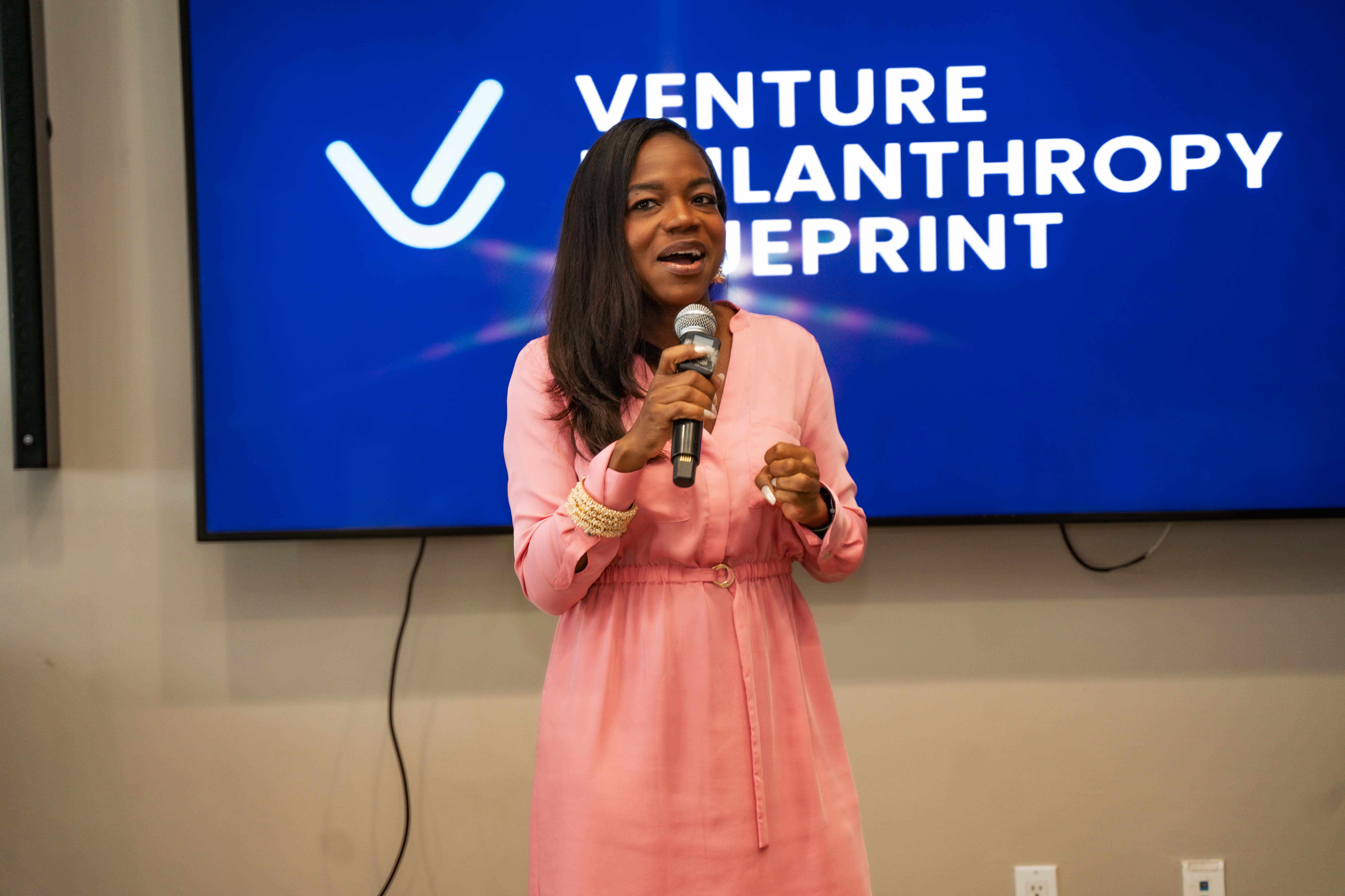 Stacey Hines, wearing a pink dress, speaks into a microphone during the Venture Philanthropy Blueprint Power Brunch event in Kingston, Jamaica. She stands in front of a large screen displaying the event’s branding, engaging the audience in a dynamic and professional setting focused on social impact and innovation.