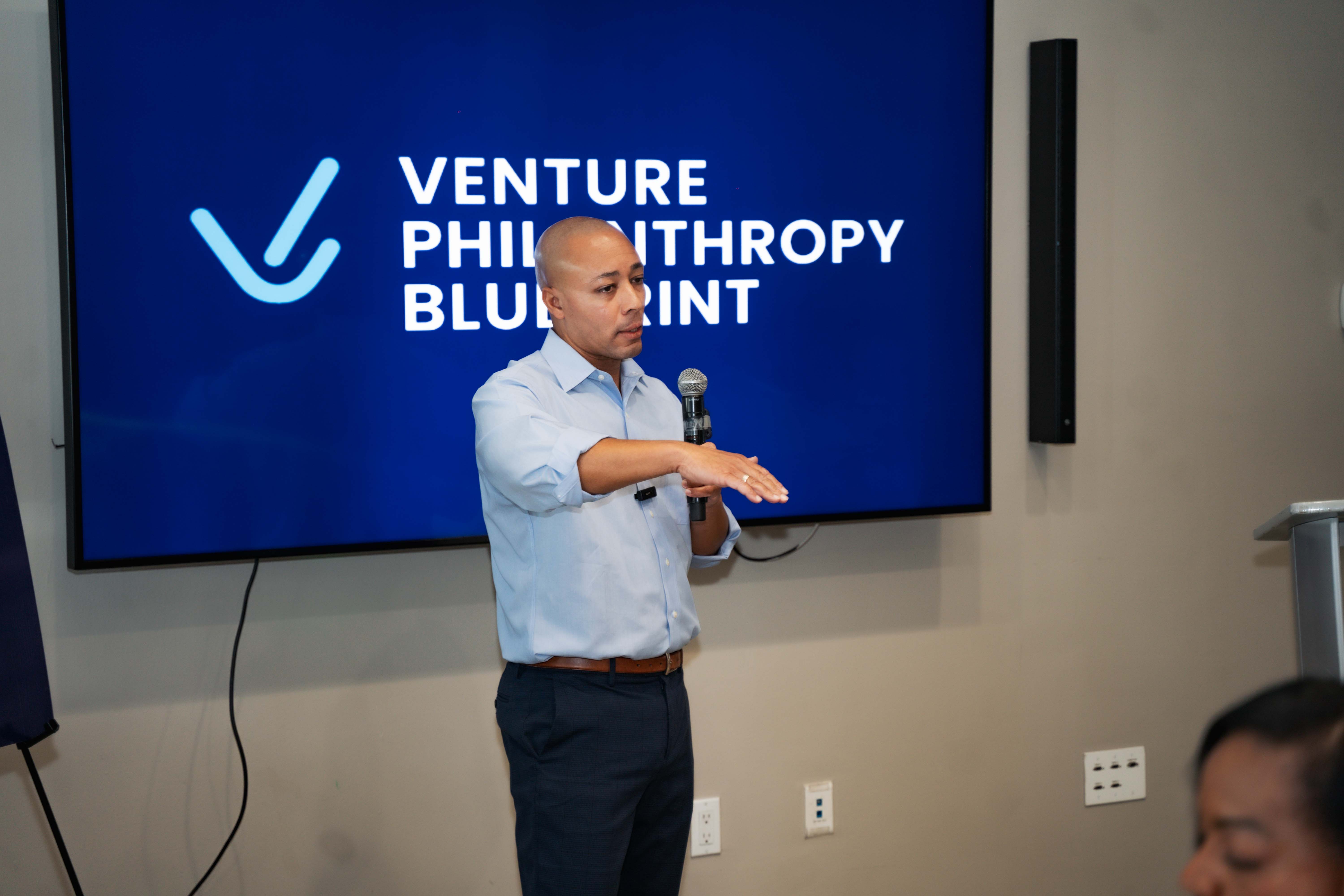 Alain Leroy, host of the Venture Philanthropy Blueprint Power Brunch event in Kingston, Jamaica, speaks into a microphone while gesturing to the audience. He stands in front of a large screen displaying the VPB logo, leading a session focused on funding and scaling mission-driven ventures.