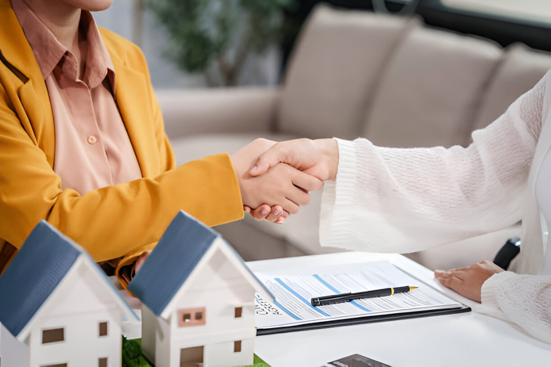 Two women shake hands over house models and a contract, representing KBR Investing helping Traditional home buyers. Two women shake hands over house models and a contract, representing KBR Investing helping Traditional home buyers.