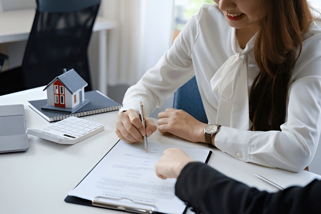A woman signs paperwork with a calculator and model house nearby, illustrating Easy home financing options through KBR Investing.
