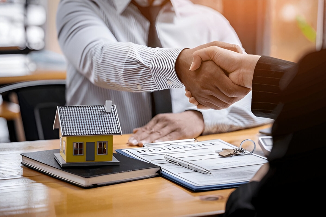 Two people shake hands over a table with a house model and documents, representing KBR Investing helping you Buy a house with low credit in the U.S.