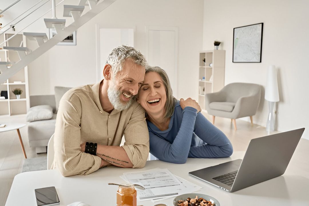 An older couple laughs while looking at a laptop at their bright kitchen table, hinting at the joy of securing Owner finance properties with no credit check through KBR Investing.