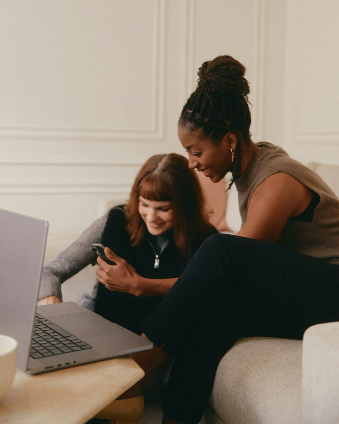 two women sitting at a computer looking at the same content on a smart phone two women sitting at a computer looking at the same content on a smart phone