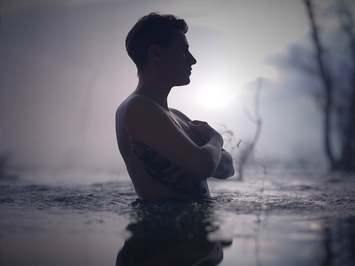 A dramatic artistic portrait showcasing a person contemplating their reflection, symbolizing introspection and confronting the past. The subject stands facing a dark, reflective pool of water, their silhouette starkly contrasting against the muted background. The scene is bathed in soft, ethereal lighting, emphasizing the shadows and creating a melancholic atmosphere that evokes a sense of quiet reflection. Wisps of fog gently rise from the water's surface, enhancing the overall sense of mystery and introspection.