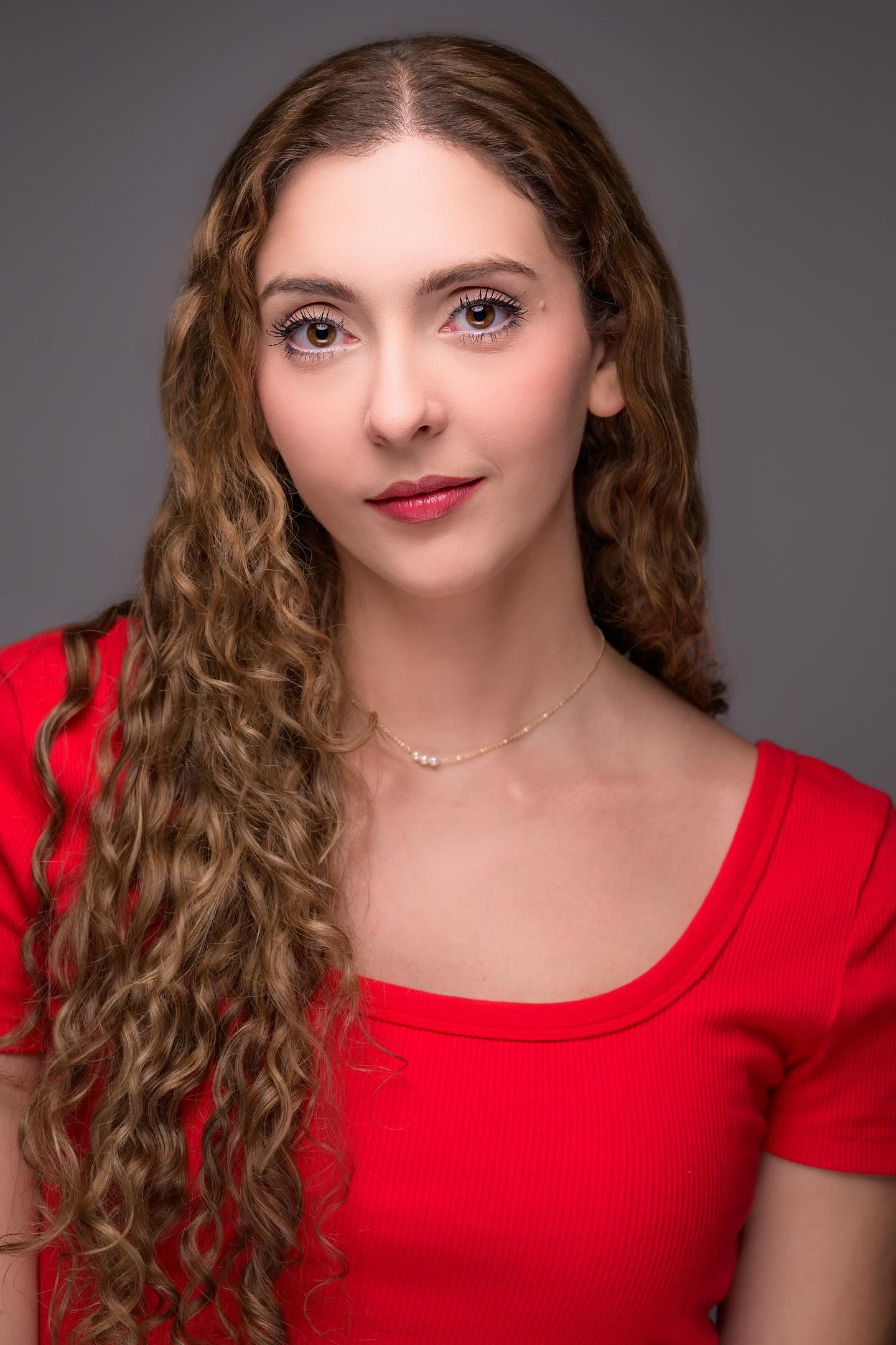 Portrait of Dana Abruzzo (@Danaabruzzo) wearing a fitted red top with a square neckline. She has long, light brown curly hair cascading over her shoulder, natural makeup with defined lashes, and soft red lipstick. She wears a delicate gold necklace with small pearls. The background is a neutral grey, and the lighting is soft and flattering, creating a professional and polished look. Portrait of Dana Abruzzo (@Danaabruzzo) wearing a fitted red top with a square neckline. She has long, light brown curly hair cascading over her shoulder, natural makeup with defined lashes, and soft red lipstick. She wears a delicate gold necklace with small pearls. The background is a neutral grey, and the lighting is soft and flattering, creating a professional and polished look.