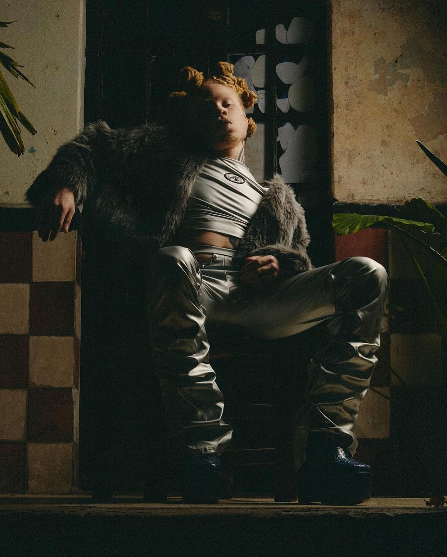 Editorial portrait of Jerrbul seated confidently on a wooden chair in a moody, dimly lit space with vintage tiled walls. He wears a silver metallic outfit featuring a crop top and matching pants, paired with a textured faux fur coat and chunky platform boots. His hair is styled in twisted bantu knots, and his relaxed, commanding pose exudes individuality and high-fashion edge. Green plants frame the composition, enhancing the raw, artistic atmosphere. Editorial portrait of Jerrbul seated confidently on a wooden chair in a moody, dimly lit space with vintage tiled walls. He wears a silver metallic outfit featuring a crop top and matching pants, paired with a textured faux fur coat and chunky platform boots. His hair is styled in twisted bantu knots, and his relaxed, commanding pose exudes individuality and high-fashion edge. Green plants frame the composition, enhancing the raw, artistic atmosphere.