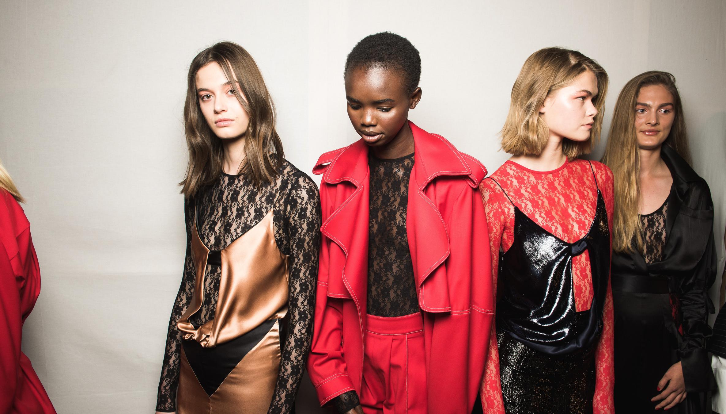 A group of five fashion models stand in a line against a neutral background, wearing stylish and modern outfits. The clothing features a mix of lace, satin, and glossy materials in bold colors such as red, black, and bronze. Each model has a distinct look, with varied hairstyles and expressions, showcasing a high-fashion aesthetic. A group of five fashion models stand in a line against a neutral background, wearing stylish and modern outfits. The clothing features a mix of lace, satin, and glossy materials in bold colors such as red, black, and bronze. Each model has a distinct look, with varied hairstyles and expressions, showcasing a high-fashion aesthetic.