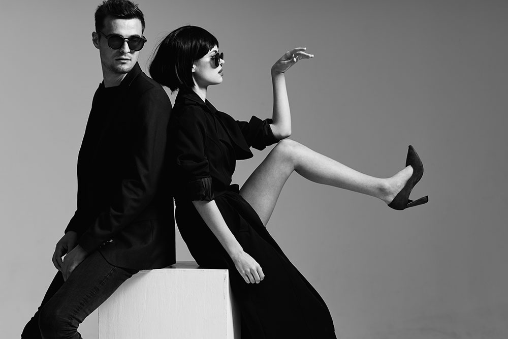 A stylish man and woman pose in a high-fashion black-and-white photo. Both are dressed in sleek black outfits and wearing dark sunglasses. The man sits on a white cube facing slightly left, while the woman leans back against him, one leg lifted dramatically and her arm raised in an elegant gesture. The scene exudes modern sophistication and minimalism. A stylish man and woman pose in a high-fashion black-and-white photo. Both are dressed in sleek black outfits and wearing dark sunglasses. The man sits on a white cube facing slightly left, while the woman leans back against him, one leg lifted dramatically and her arm raised in an elegant gesture. The scene exudes modern sophistication and minimalism.