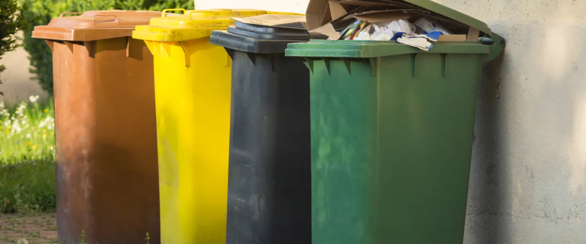 Set of four overflowing trash bins in brown, yellow, black, and green, placed beside a building