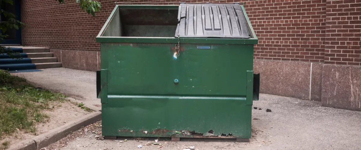 Worn green dumpster with rust and peeling paint placed in a narrow alleyway next to a brick wall