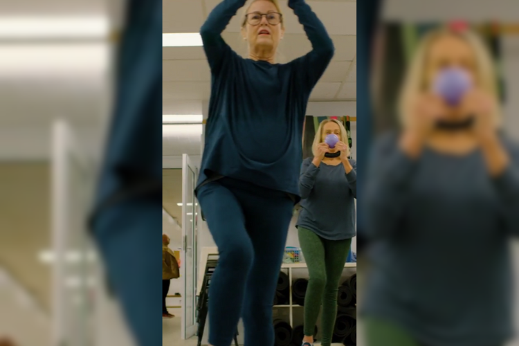 Catherine, a member of Community Moves Health & Fitness, participates in a group exercise class. She lifts her arms above her head, showing focus and strength. Another woman stands behind her, holding a small exercise ball, highlighting the supportive and engaging environment of Community Moves' fitness programs for adults over 50. Catherine, a member of Community Moves Health & Fitness, participates in a group exercise class. She lifts her arms above her head, showing focus and strength. Another woman stands behind her, holding a small exercise ball, highlighting the supportive and engaging environment of Community Moves' fitness programs for adults over 50.