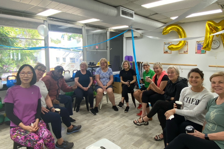 A group of Community Moves Health & Fitness members, predominantly women over 50, gathered in the gym for a social event, sitting in a circle with coffee cups, smiling and enjoying each other's company. The room is decorated with blue streamers and gold balloons, creating a festive atmosphere. A group of Community Moves Health & Fitness members, predominantly women over 50, gathered in the gym for a social event, sitting in a circle with coffee cups, smiling and enjoying each other's company. The room is decorated with blue streamers and gold balloons, creating a festive atmosphere.