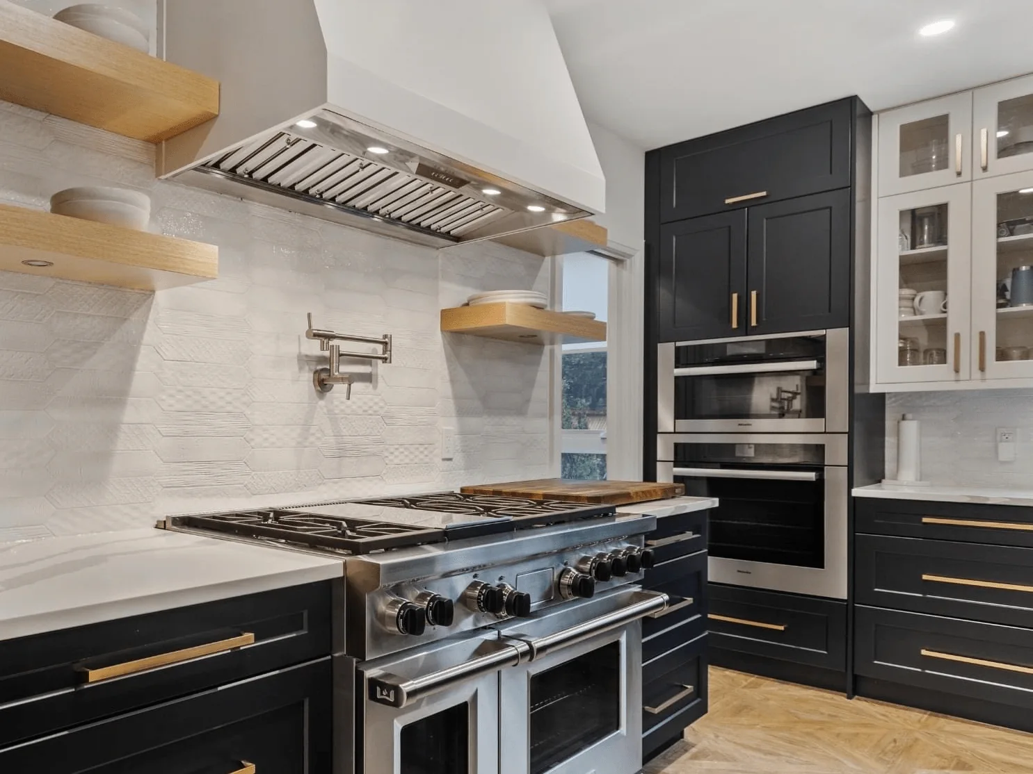 A modern kitchen with painting black cabinets, a stainless steel range, and open wooden shelves against a white tile backsplash.