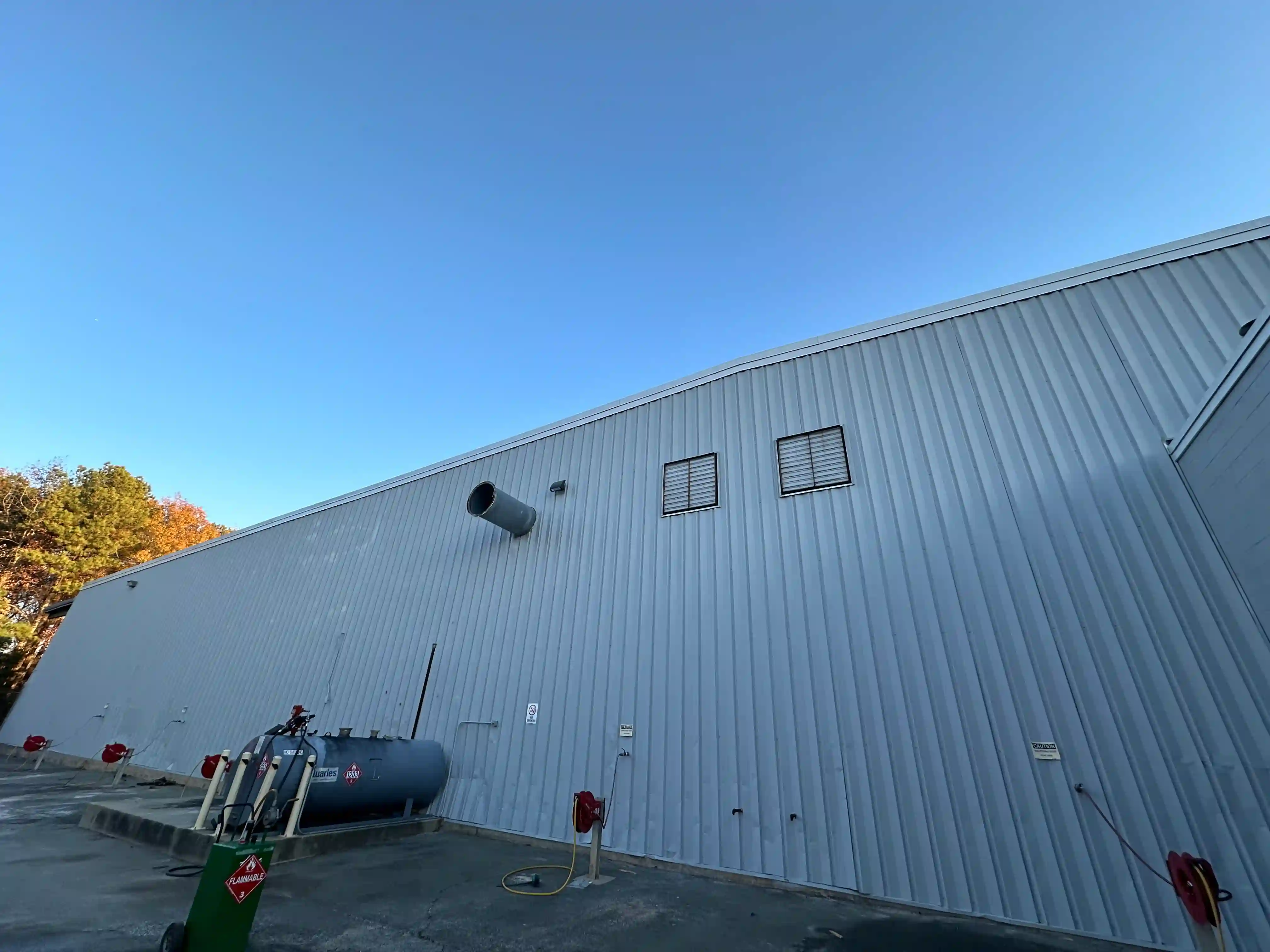 A large industrial building with a smooth, painting metal exterior, featuring a fuel tank and safety equipment in the foreground, set against a clear blue sky.