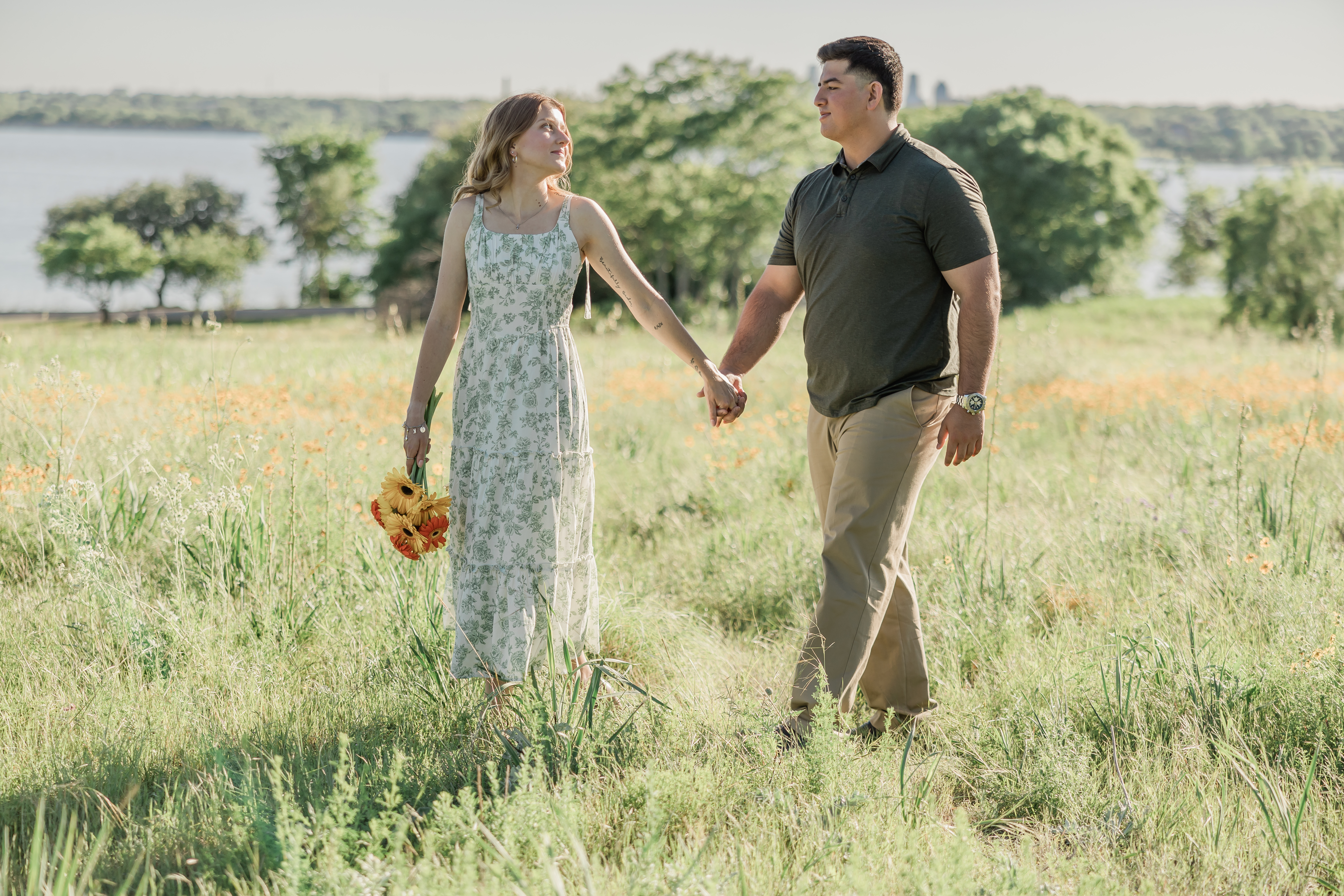 An image of an engaged couple walking in a field on Winfrey Point with White Rock Lake in the background, taken by Arlene Stepanian Photography, a wedding photographer in Dallas.