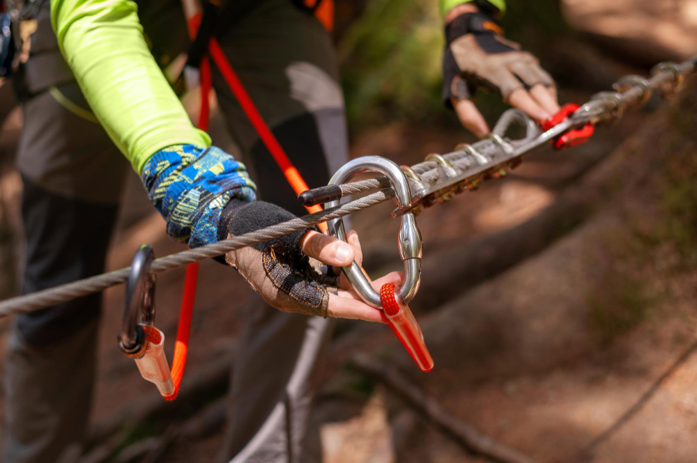 Close-up of an arborist’s gloved hand adjusting a carabiner on a metal safety cable. The person wears a long-sleeve shirt and is engaged in a tree climbing or high-angle rigging activity, emphasizing safety procedures in arboriculture.