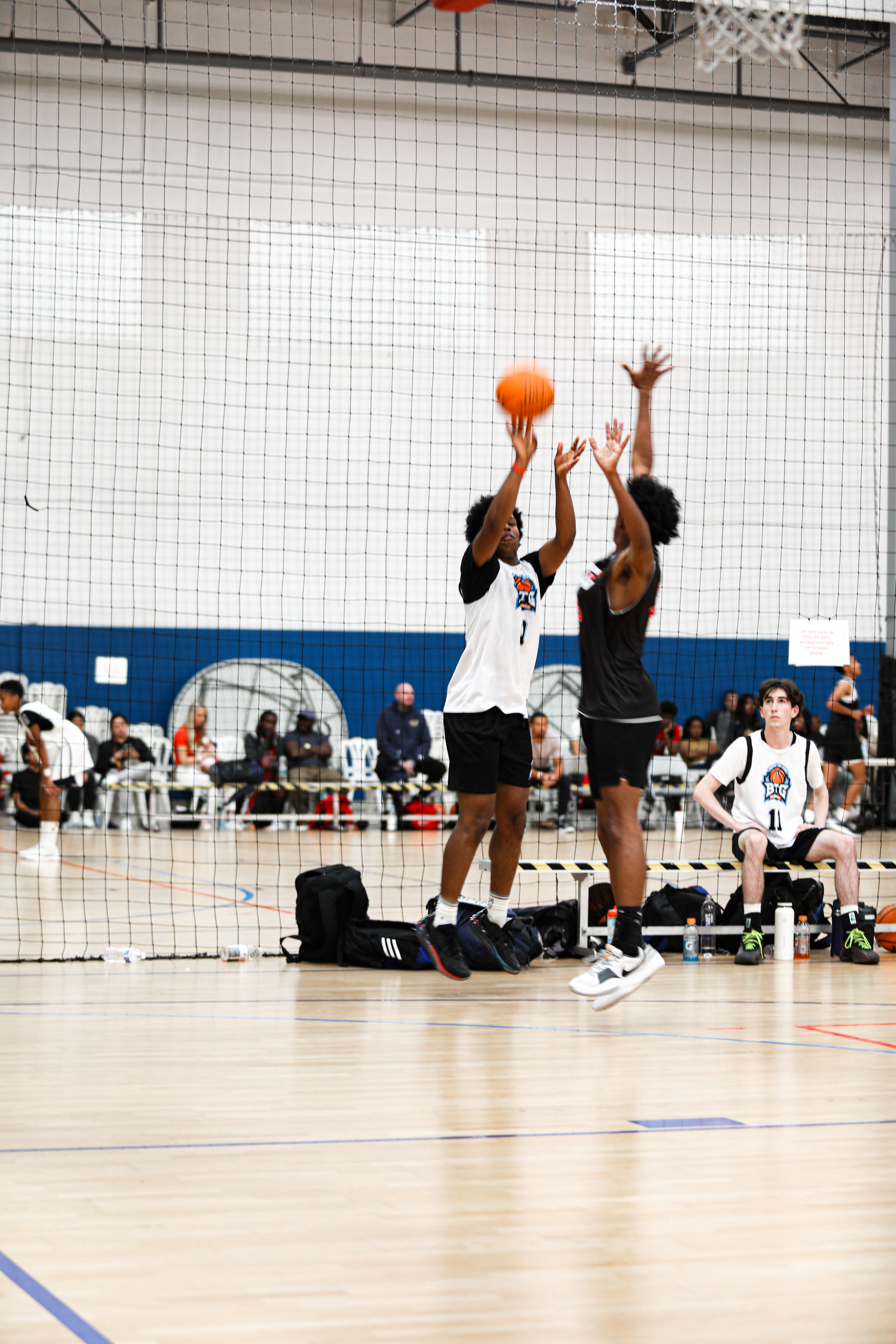 High school male basketball player shooting a basketball while contested by another player in an indoor court