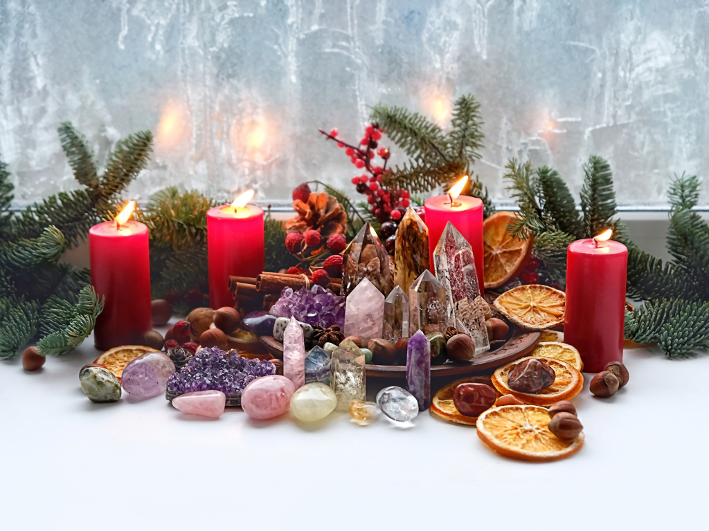 Lit red candles are surrounded by pine branches, crystals, and dried oranges. Lit red candles are surrounded by pine branches, crystals, and dried oranges.