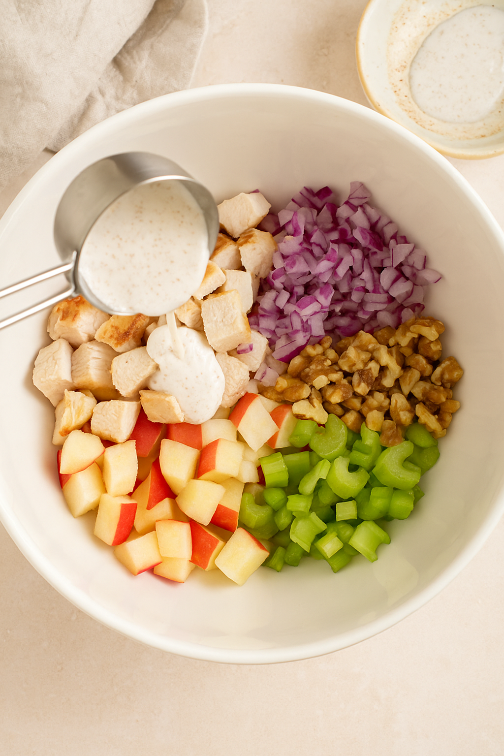 Pouring creamy coconut yogurt dressing over diced chicken, apples, celery, red onion, and walnuts in a mixing bowl for Apple-Walnut Chicken Salad.