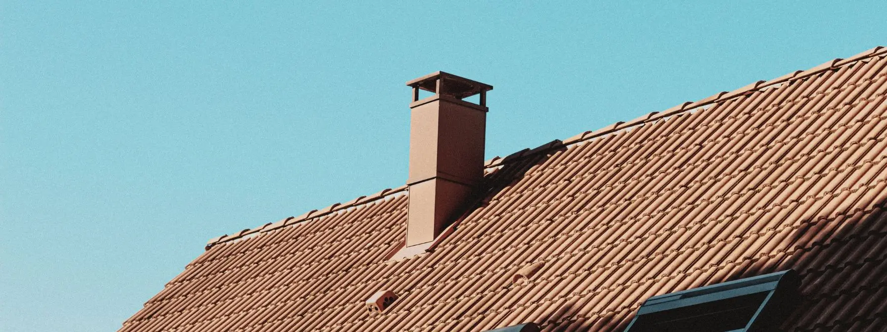 A clay tile roof with a chimney stack under a clear blue sky, illustrating how summer heat and UV exposure can affect different roofing materials in Ashland, KY. A clay tile roof with a chimney stack under a clear blue sky, illustrating how summer heat and UV exposure can affect different roofing materials in Ashland, KY.