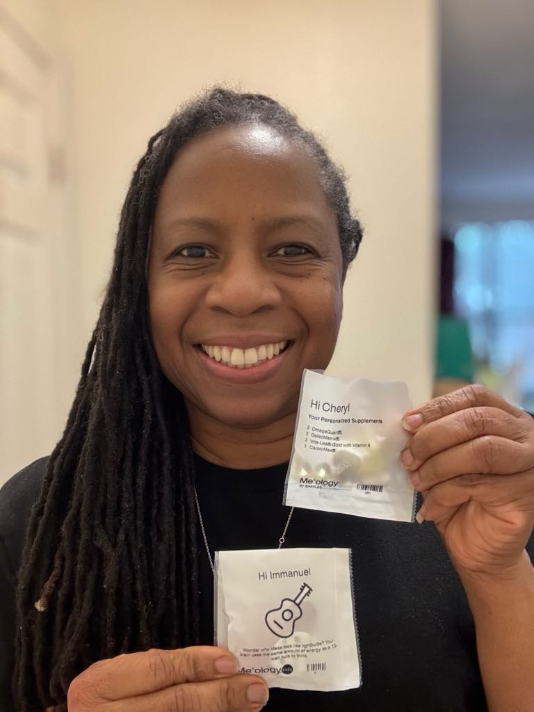 Cheryl Adams smiling and holding personalized Meology vitamin packs for herself and her son, showcasing customized daily supplements.