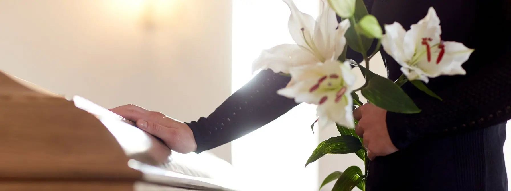 Person in mourning attire holding white lilies and placing a hand on a wooden casket, symbolizing loss and the importance of having a will to guide estate decisions in West Virginia. Person in mourning attire holding white lilies and placing a hand on a wooden casket, symbolizing loss and the importance of having a will to guide estate decisions in West Virginia.