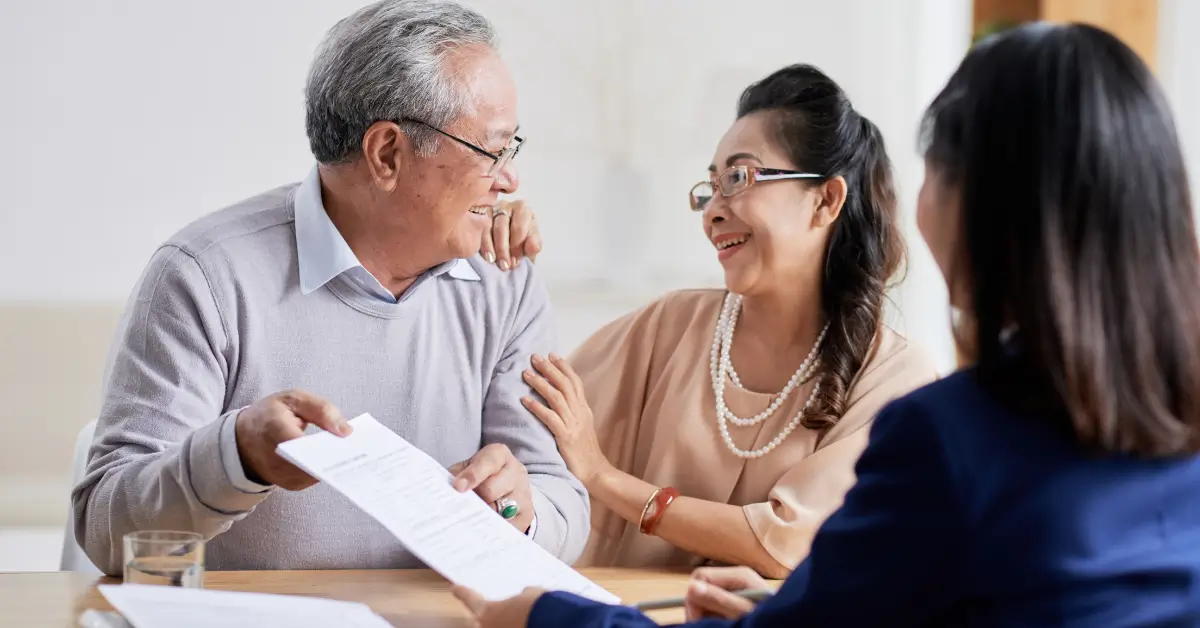 An elderly couple smiling and holding documents while sitting across from a professional, possibly discussing estate planning or legal matters, in a bright indoor setting. An elderly couple smiling and holding documents while sitting across from a professional, possibly discussing estate planning or legal matters, in a bright indoor setting.