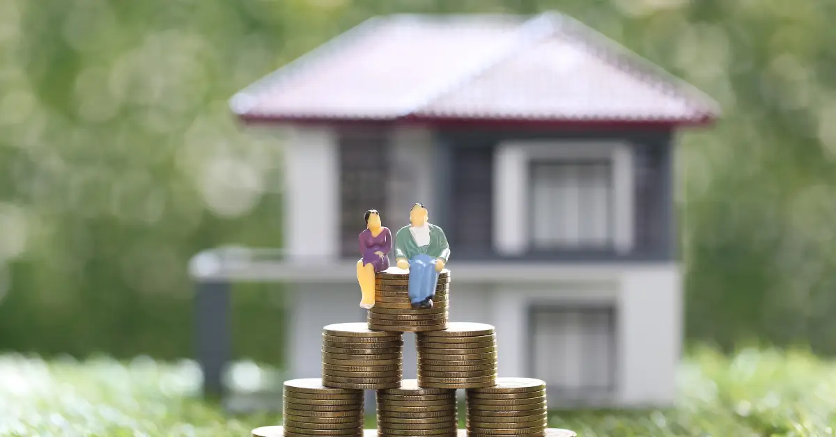 Miniature figurines of a couple sitting on top of stacked coins with a blurred model house in the background, symbolizing real estate investment, savings, or retirement planning. Miniature figurines of a couple sitting on top of stacked coins with a blurred model house in the background, symbolizing real estate investment, savings, or retirement planning.