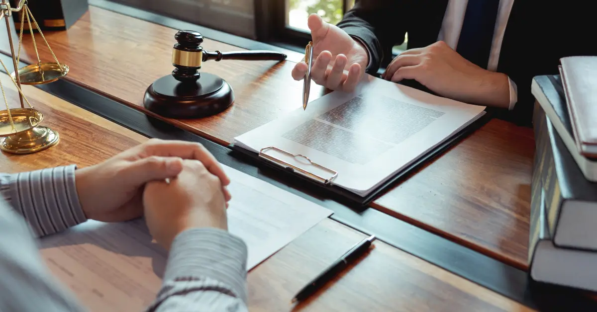 Close-up of a legal consultation, showing a lawyer pointing at a document on a clipboard while another person listens, with a gavel, scales of justice, and law books on the desk. Close-up of a legal consultation, showing a lawyer pointing at a document on a clipboard while another person listens, with a gavel, scales of justice, and law books on the desk.