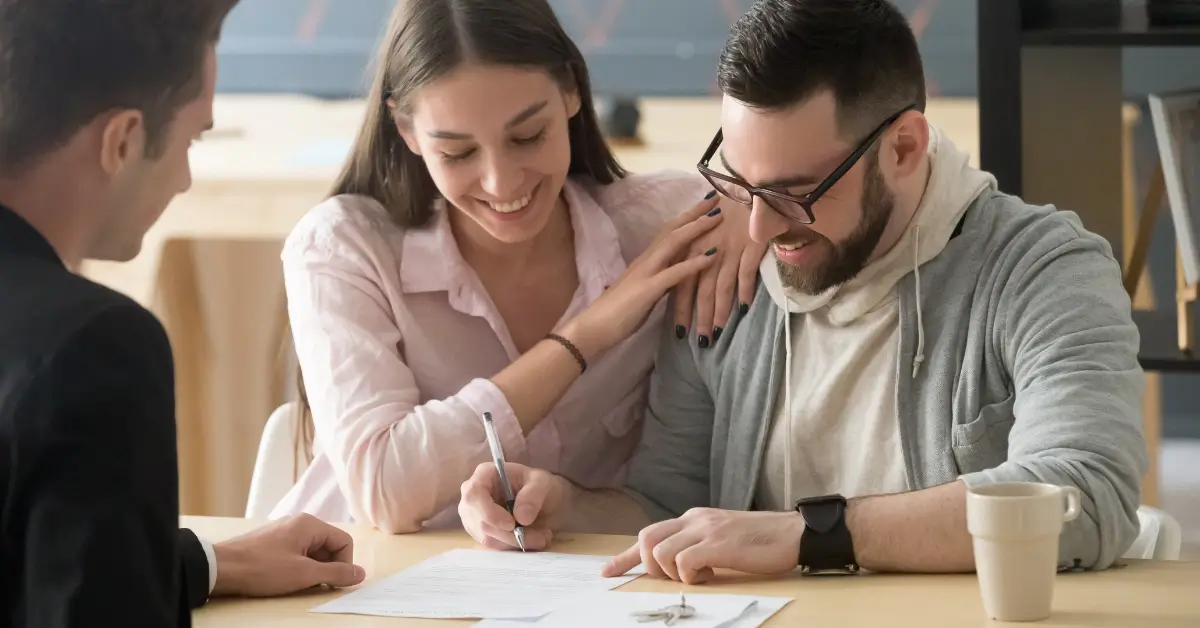 Smiling couple sitting together at a table, with the woman signing a document while the man looks on, and a professional sitting across from them.