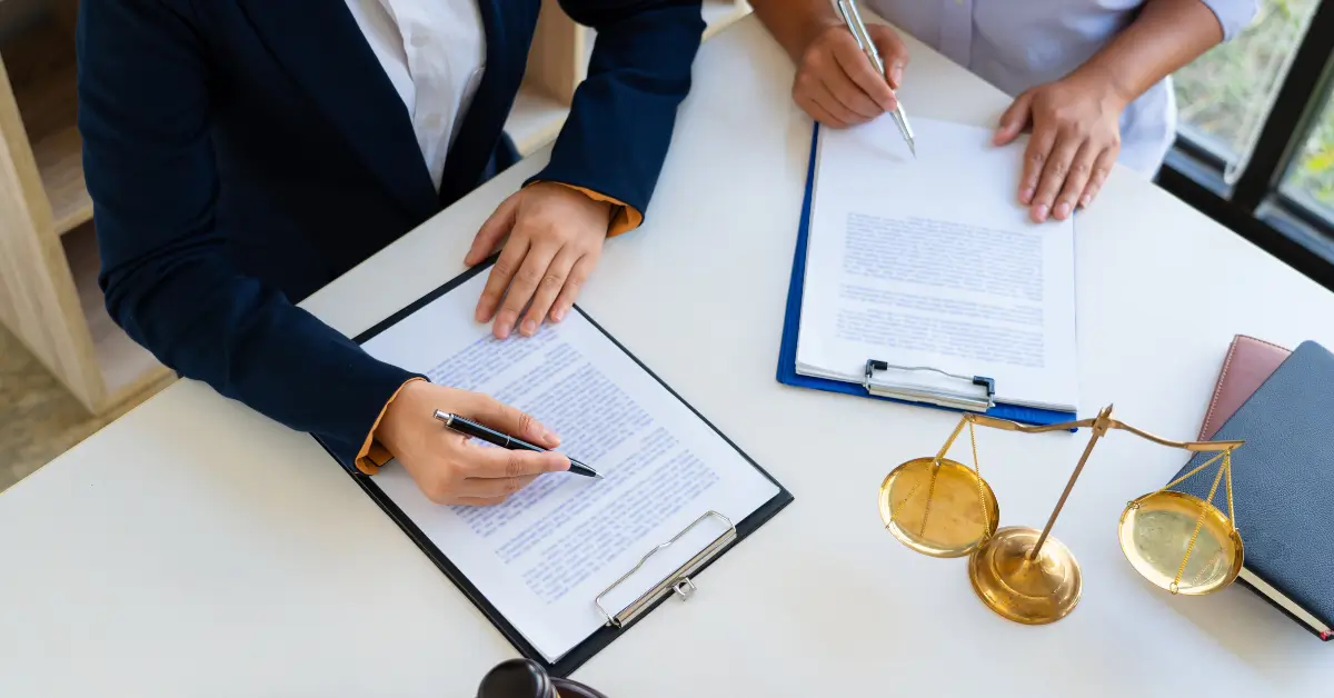 Two people sitting at a desk reviewing and signing documents on clipboards, with a golden balance scale and books on the table, symbolizing legal or contractual matters. Two people sitting at a desk reviewing and signing documents on clipboards, with a golden balance scale and books on the table, symbolizing legal or contractual matters.