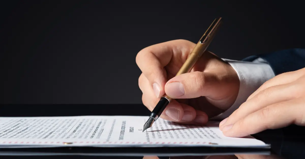 A person signing a legal document with a gold fountain pen against a dark background.