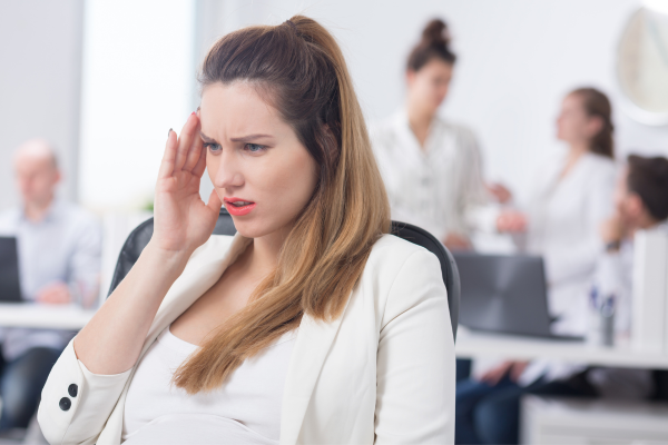 A concerned woman holding her head in discomfort while seated in a busy office, symbolizing invisible symptoms, overwhelm, and the emotional toll of being misunderstood. A concerned woman holding her head in discomfort while seated in a busy office, symbolizing invisible symptoms, overwhelm, and the emotional toll of being misunderstood.