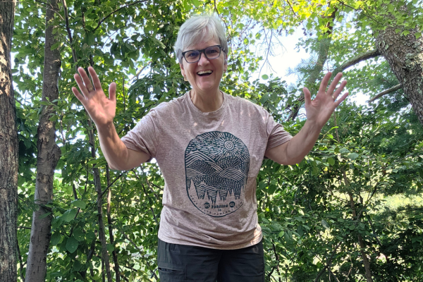 Claudette Eames smiling with arms open on a forest trail, symbolizing freedom and building a business that fits your life.