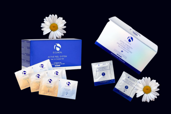 Two IS CLINICAL Active Peel System skincare boxes displayed on a dark background, each with individual peel packets arranged in front. White daisy flowers are placed near both boxes for decoration. Two IS CLINICAL Active Peel System skincare boxes displayed on a dark background, each with individual peel packets arranged in front. White daisy flowers are placed near both boxes for decoration.