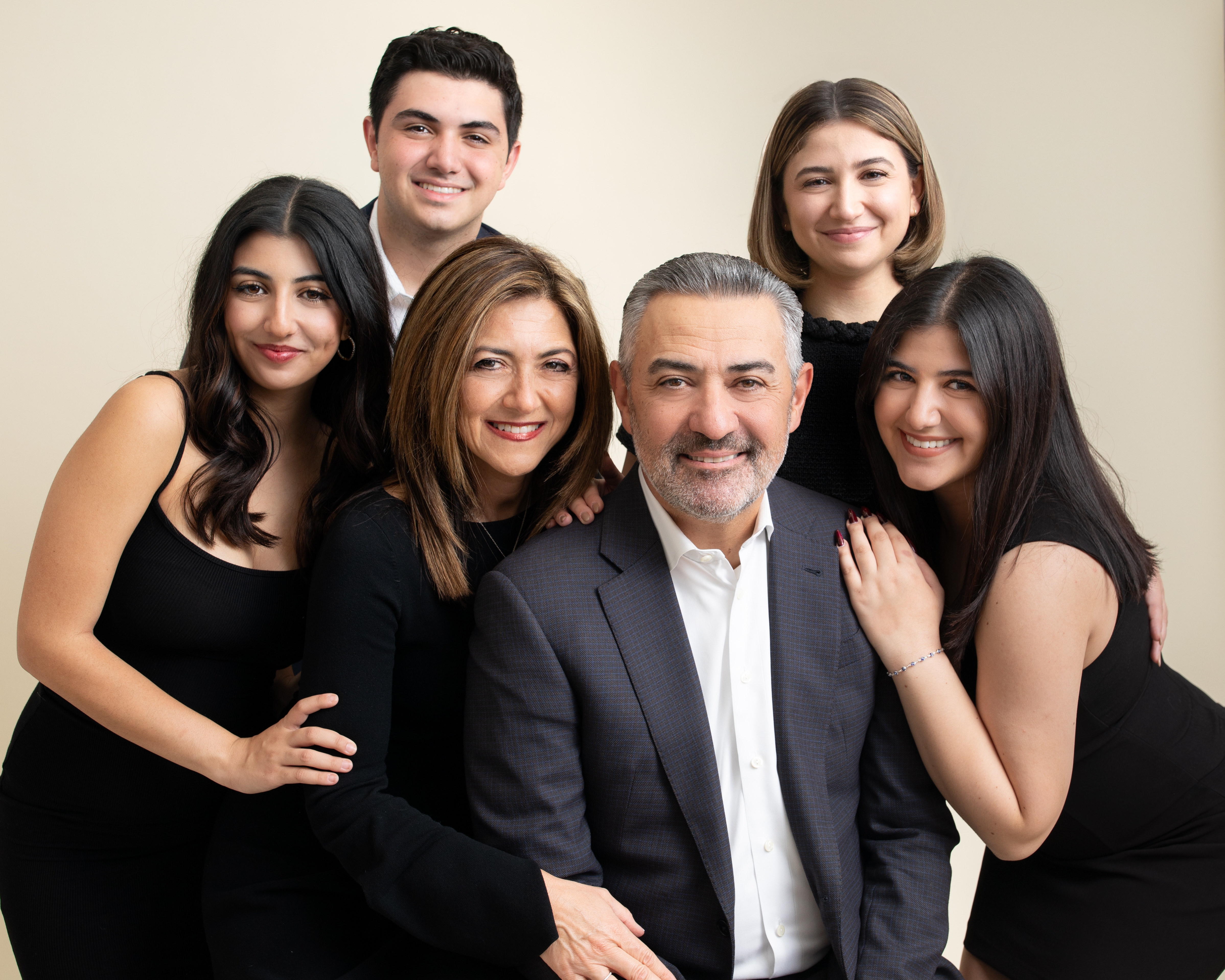 A family of six smiles warmly while dressed in classic black and white, gathered closely together in front of a light background. Photographed by Los Angeles photographer Kendra Greenberg Photography.