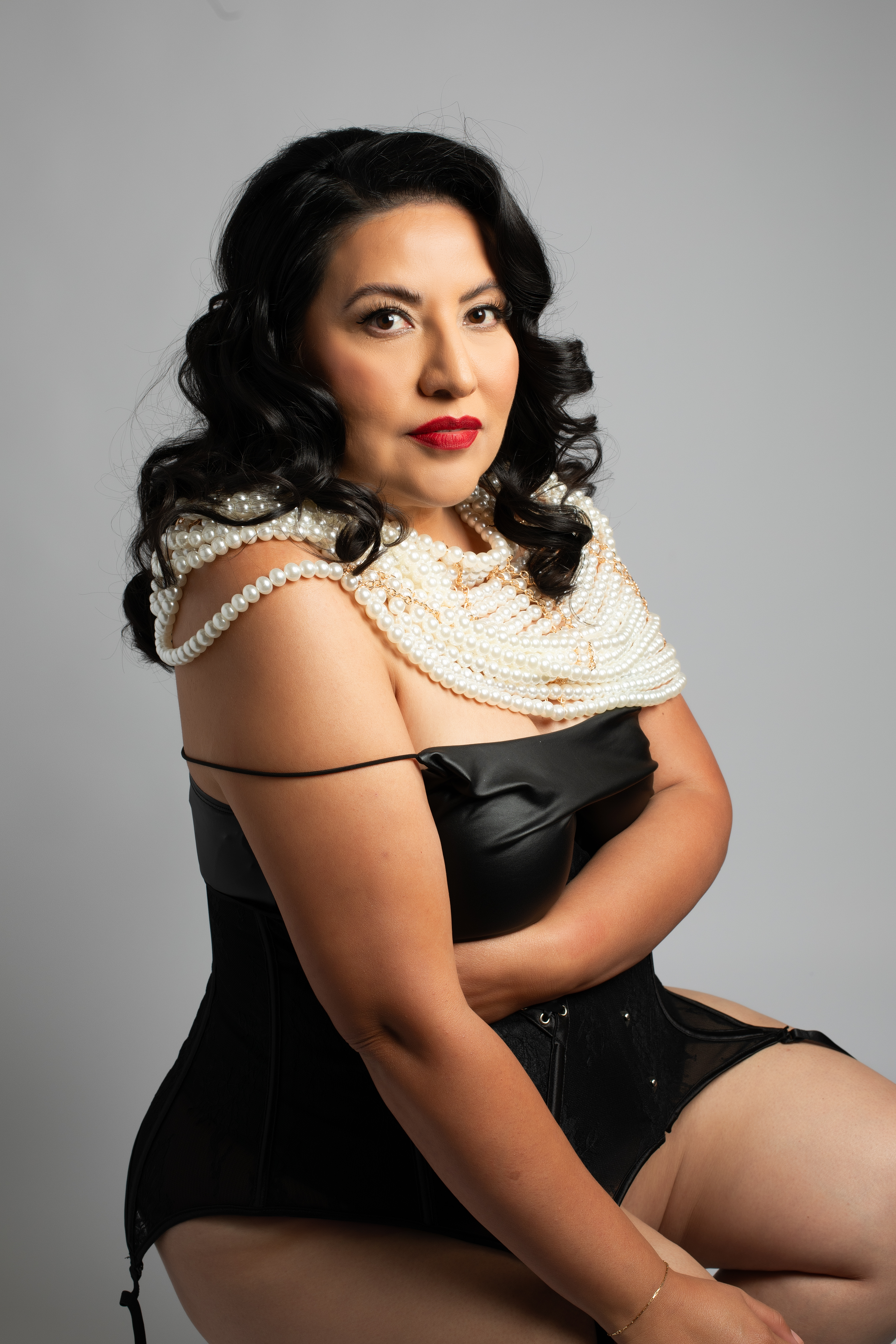 A confident woman poses against a neutral backdrop wearing a black corset and layered pearl necklace. Her dark, wavy hair frames her face as she gazes directly at the camera, exuding timeless elegance and strength. Photographed by Los Angeles boudoir photographer Kendra Greenberg Photography.
