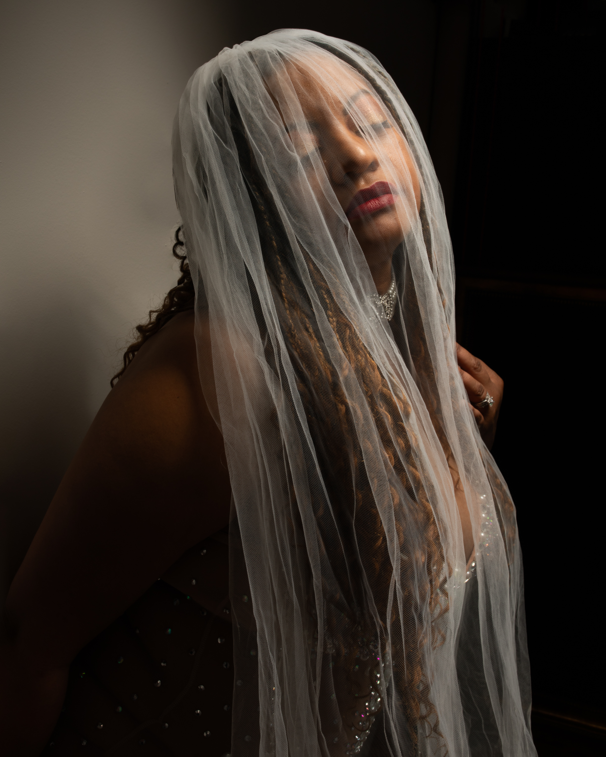 A woman stands softly lit against a dark background, her eyes closed beneath a sheer white veil that drapes over her face and shoulders. The light catches the shimmer of her jeweled dress and highlights her serene, confident expression. Photographed by Los Angeles boudoir photographer Kendra Greenberg Photography.
