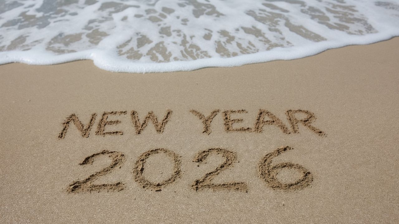 New Year 2026" written in the sand on a beach with an ocean wave approaching, symbolizing a fresh start and reflection before setting new goals.