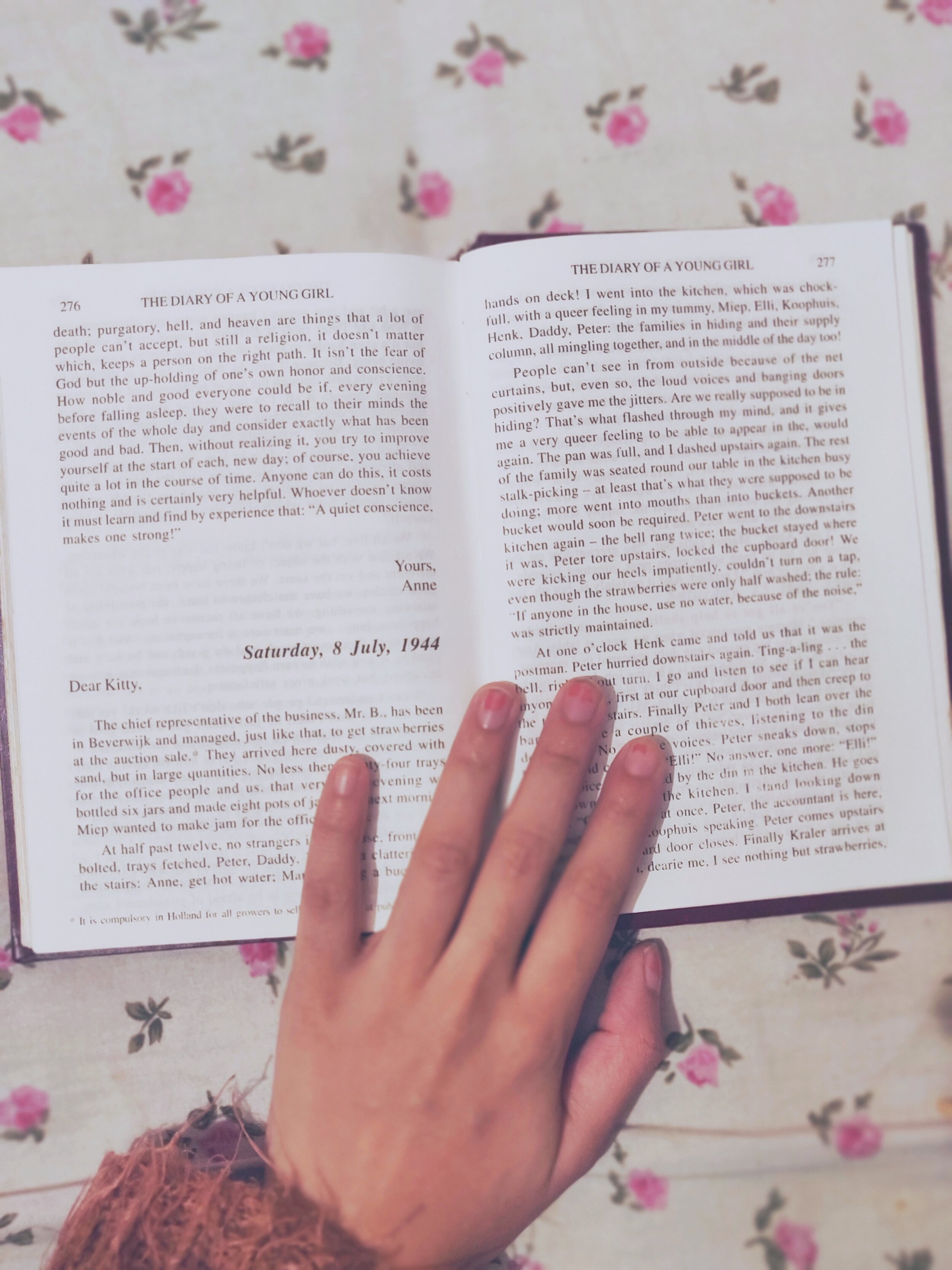  A photograph of the book 'The Diary of Anne Frank' open at the entry for Saturday 8th July 1944. A hand rests on the book and the image is set against a  print of pink rose buds on a  white background