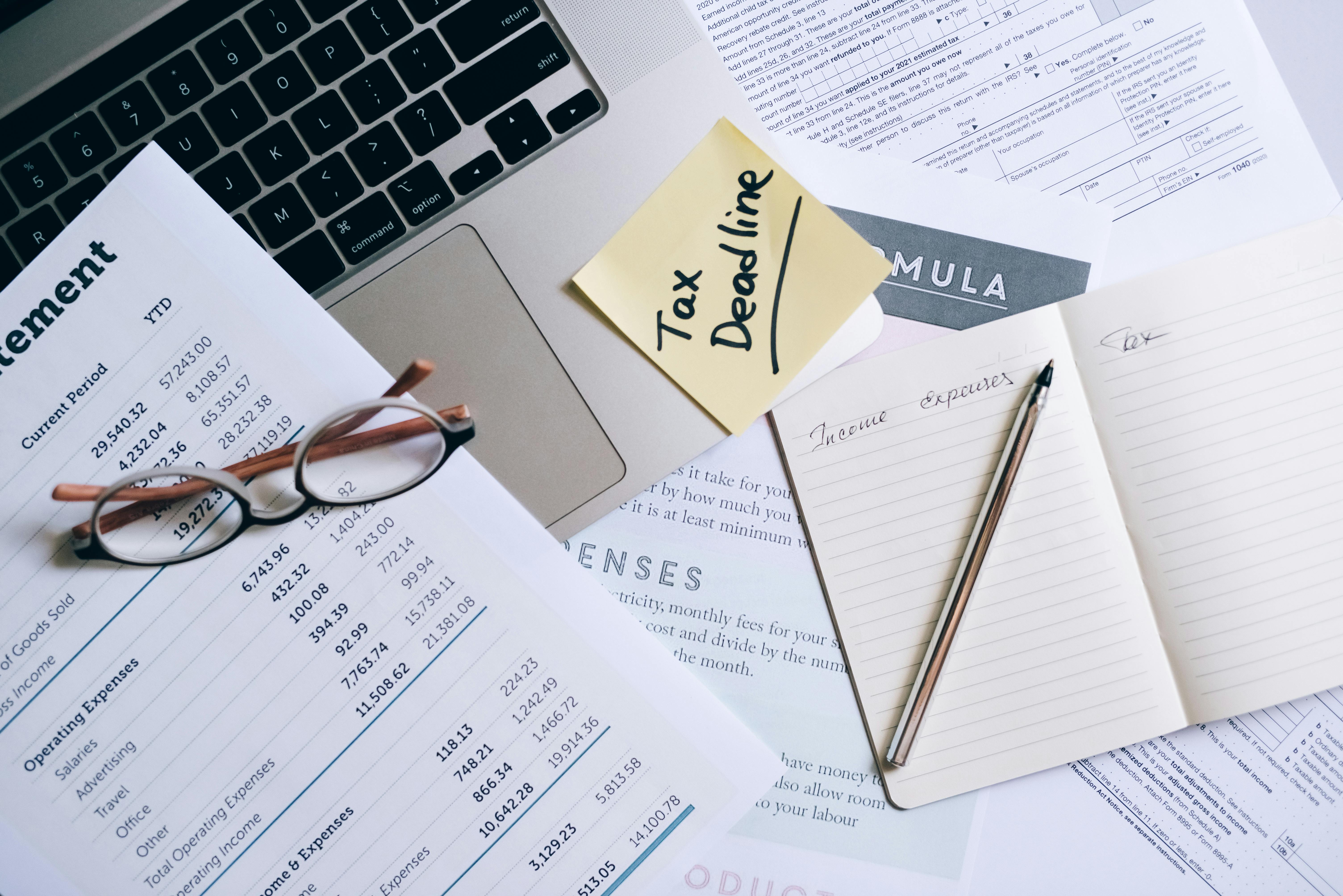 Flat lay of financial documents, income and expense sheets, a laptop, a sticky note labeled “Tax Deadline,” eyeglasses, and a notebook with handwritten notes—representing tax preparation, accounting, and small business financial planning.
