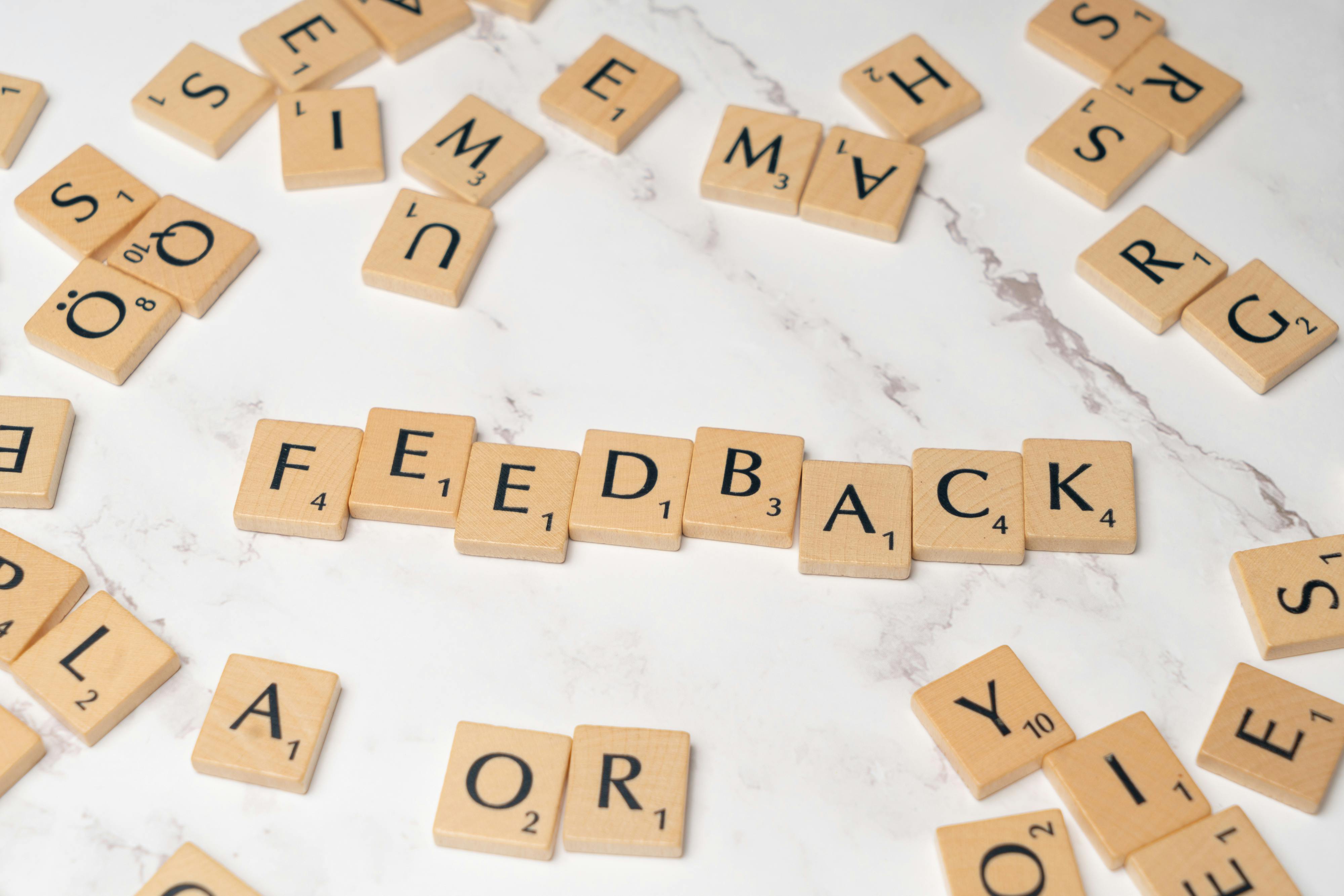Wooden letter tiles arranged to spell “FEEDBACK” on a white marble surface, surrounded by scattered tiles—representing customer feedback, user reviews, communication, and brand improvement strategies. Wooden letter tiles arranged to spell “FEEDBACK” on a white marble surface, surrounded by scattered tiles—representing customer feedback, user reviews, communication, and brand improvement strategies.