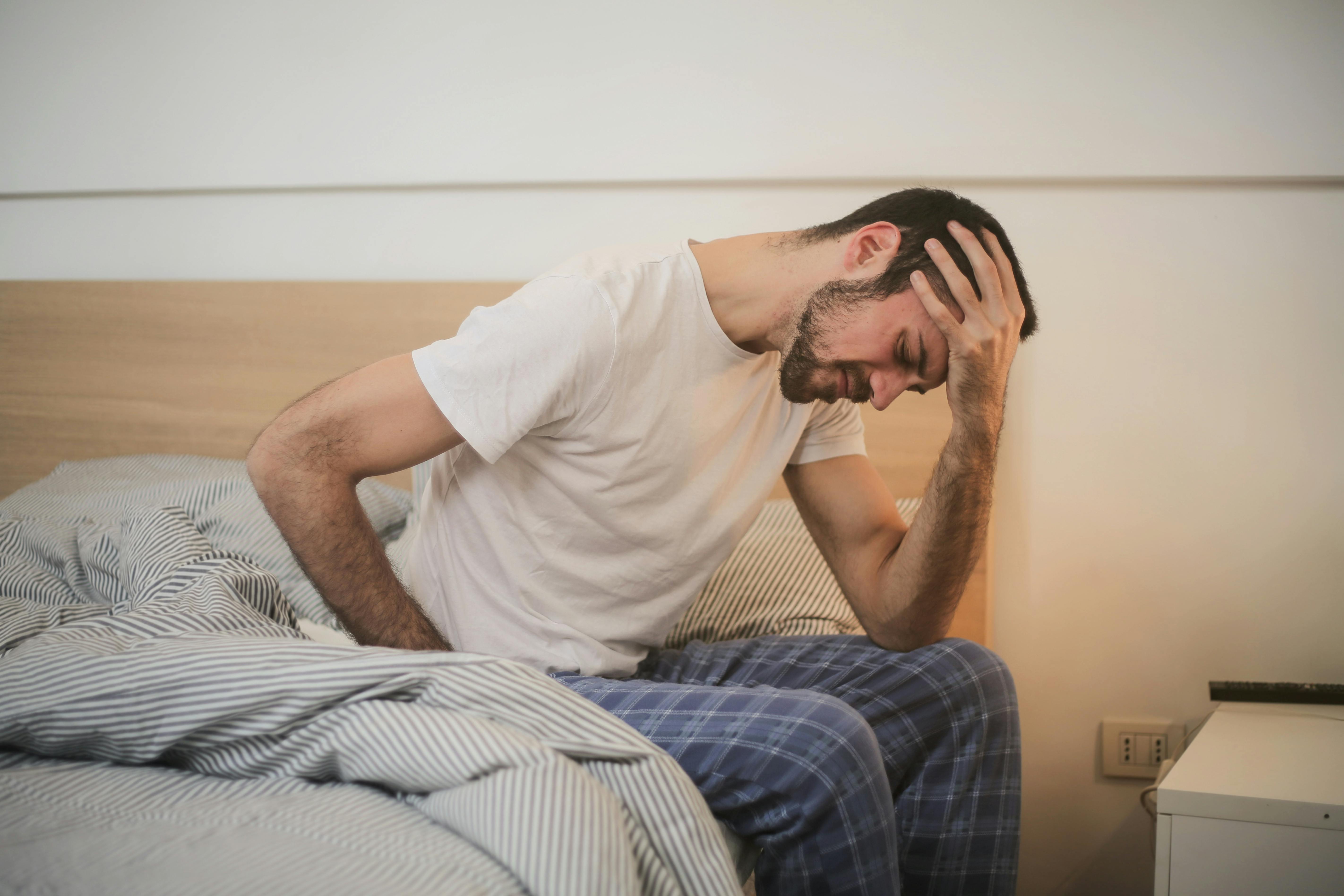 Man sitting on bed holding his head with pain, showing headache and fatigue linked to mould exposure at home.