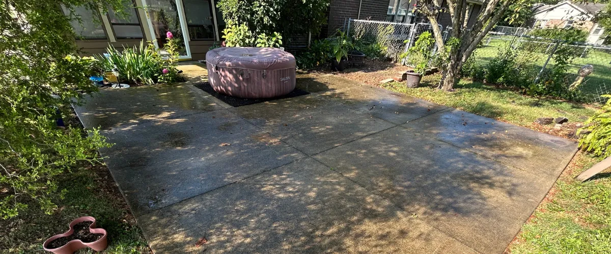Concrete patio showing wet sections after pressure washing, with plants, a hot tub, and yard space in the background.