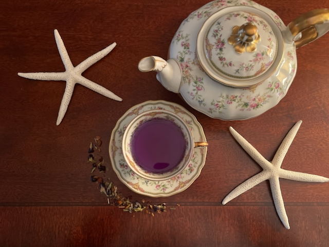 Set on dark red wooden side table, a Caribbean feel, Harney & Sons Indigo Punch has been poured in a porcelain teacup. Tropical dried herbal tea hugs the saucer, sprinkled in an arc, two decorative starfish and a porcelain teapot provide a feeling of travel and luxury. Set on dark red wooden side table, a Caribbean feel, Harney & Sons Indigo Punch has been poured in a porcelain teacup. Tropical dried herbal tea hugs the saucer, sprinkled in an arc, two decorative starfish and a porcelain teapot provide a feeling of travel and luxury.