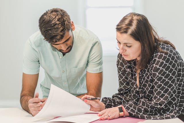 tw people checking a document a woman siting down look down at the document and a man banding over her sholder look at it also