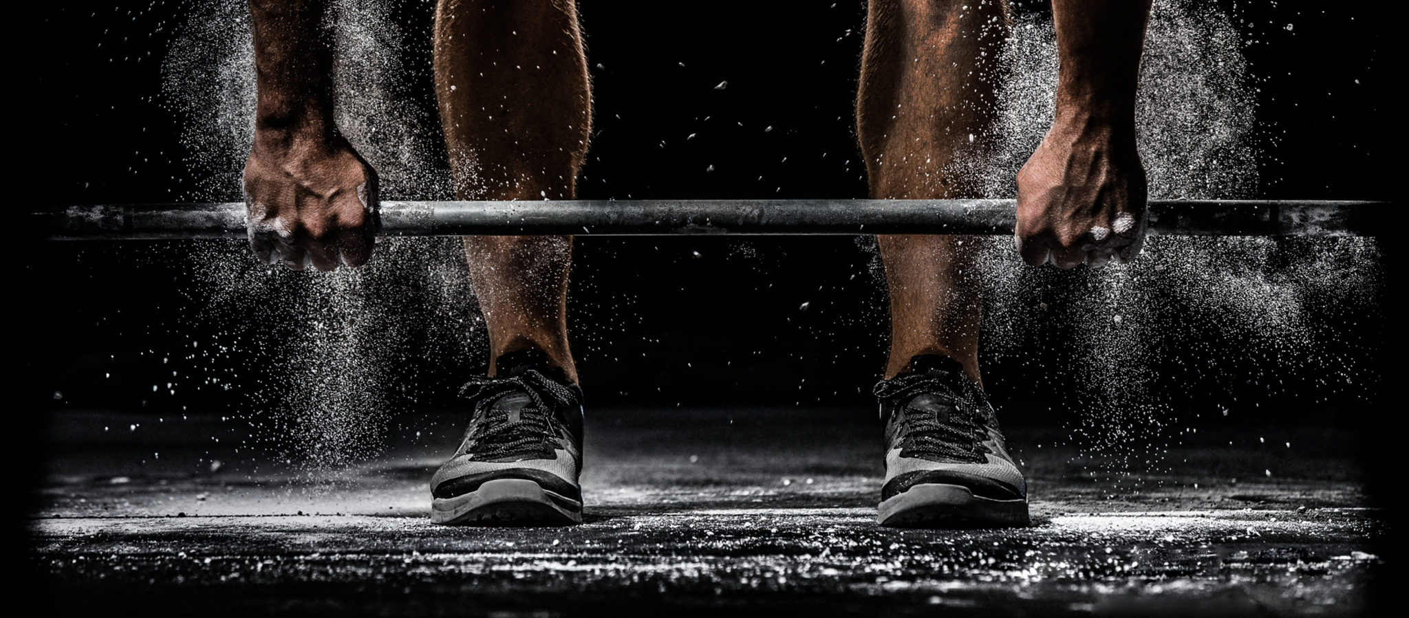 Close up picture of man's feet and legs while grabbing a bar to execute a deadlift with chalk falling.