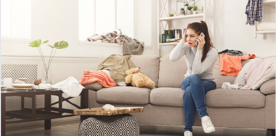 A young woman sitting on a beige sofa in a cluttered living room, holding a phone to her ear with a worried expression. The space is filled with scattered clothes, an unkempt coffee table with a pizza box, and a messy environment, conveying a sense of stress and disorganization. A young woman sitting on a beige sofa in a cluttered living room, holding a phone to her ear with a worried expression. The space is filled with scattered clothes, an unkempt coffee table with a pizza box, and a messy environment, conveying a sense of stress and disorganization.